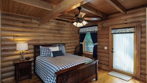 A log cabin bedroom with a wooden bed, a nightstand with a lamp, a ceiling fan, and a window with curtains. - Peaceful Paradise secluded mountain log cabin with scenic view and game room (Whittier)