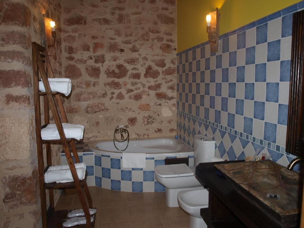 Un baño con pared de piedra, salpicadero de baldosas de cuadros, toallero de madera, bañera, inodoro y lavabo.