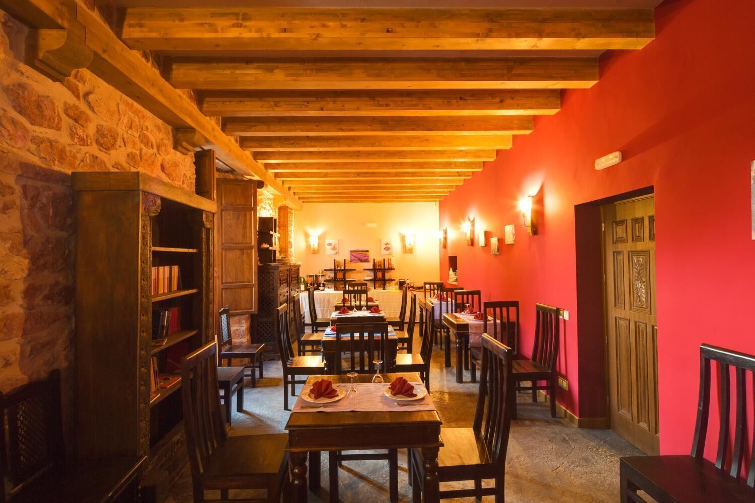 A cozy restaurant with wooden beams, stone walls, and red accents. Tables are set with plates and glasses.