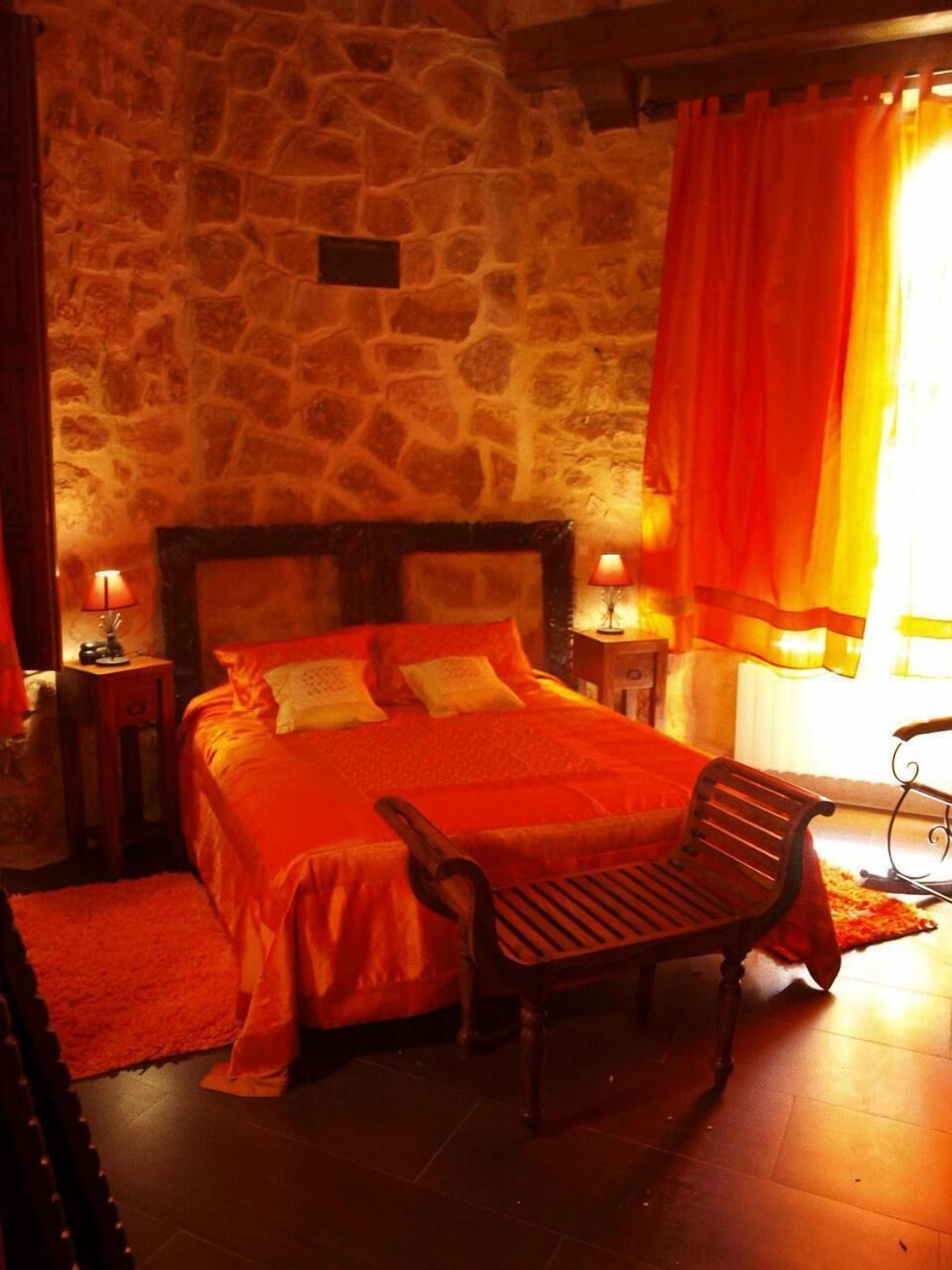 Un dormitorio con paredes de piedra, una cama grande, dos mesitas de noche con lámparas, un banco de madera y una ventana con cortinas naranjas.