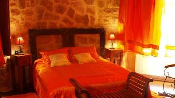 Un dormitorio con paredes de piedra, una cama grande, dos mesitas de noche con lámparas, un banco de madera y una ventana con cortinas naranjas.
