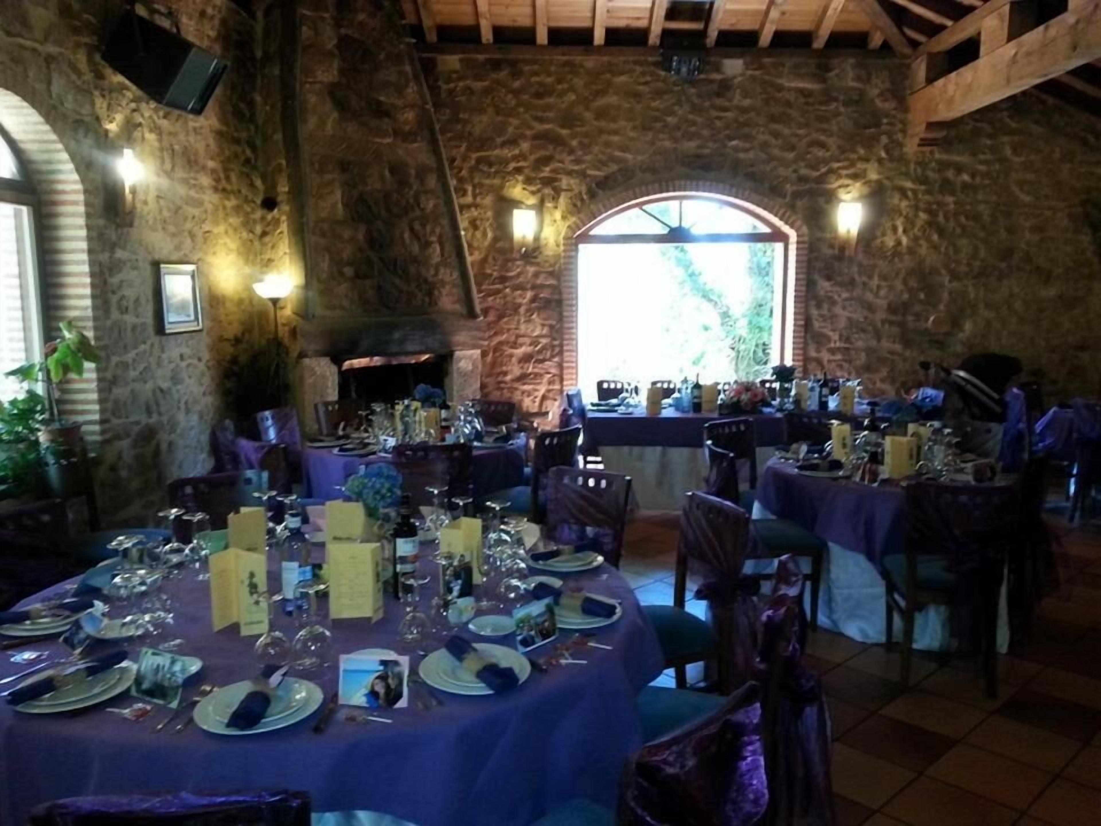 Un comedor rústico con paredes de piedra, chimenea y vistas a través de una ventana arqueada. Las mesas están preparadas con platos, vasos y cubiertos.