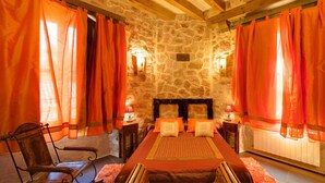 A stone-walled bedroom with a bed, two nightstands, a chair, and orange curtains. - Posada Real El Señorío de la Serrezuela (Aldeanueva De La Serrezuela)