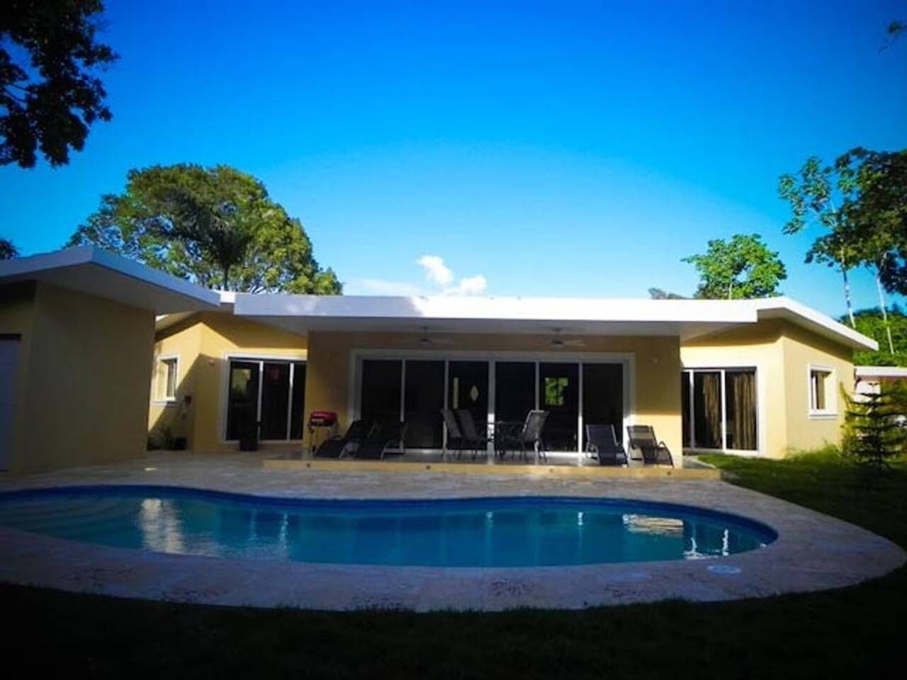 Una casa moderna con piscina, un porche cubierto con sillas y un amplio ventanal que da a la zona de la piscina.