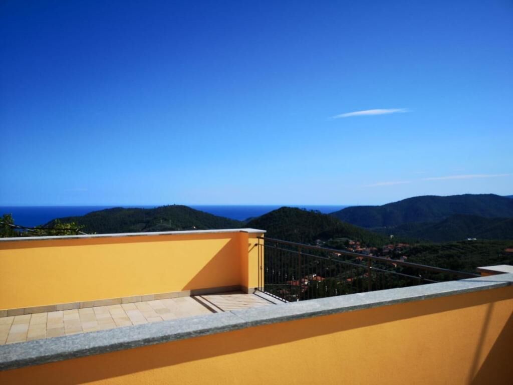 Ein gelber Balkon mit Metallgeländer, der auf eine Küstenlandschaft mit Hügeln und blauem Himmel blickt.