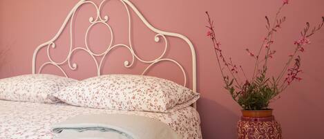 A neatly made bed with floral bedding, a decorative metal headboard, and a vase with pink flowers on a wooden stool.