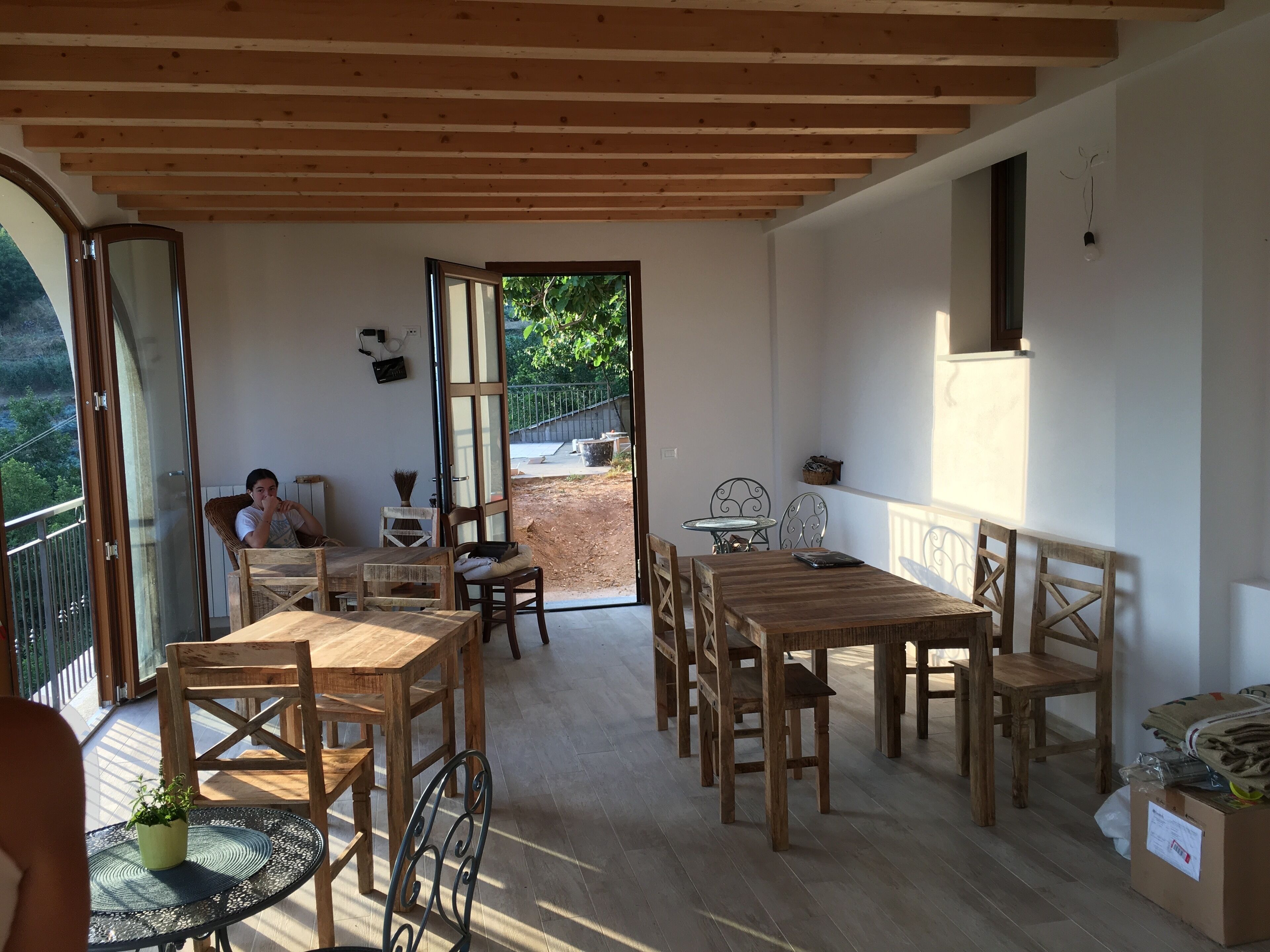 Eine geräumige Terrasse mit Holztischen und -stühlen, ein kleiner runder Tisch mit einem Blumentopf und Blick auf einen Garten durch eine offene Tür.