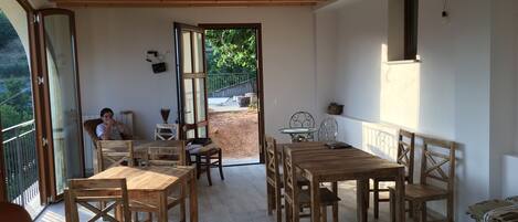 A spacious patio with wooden tables and chairs, a small round table with a potted plant, and a view of a garden through an open door.