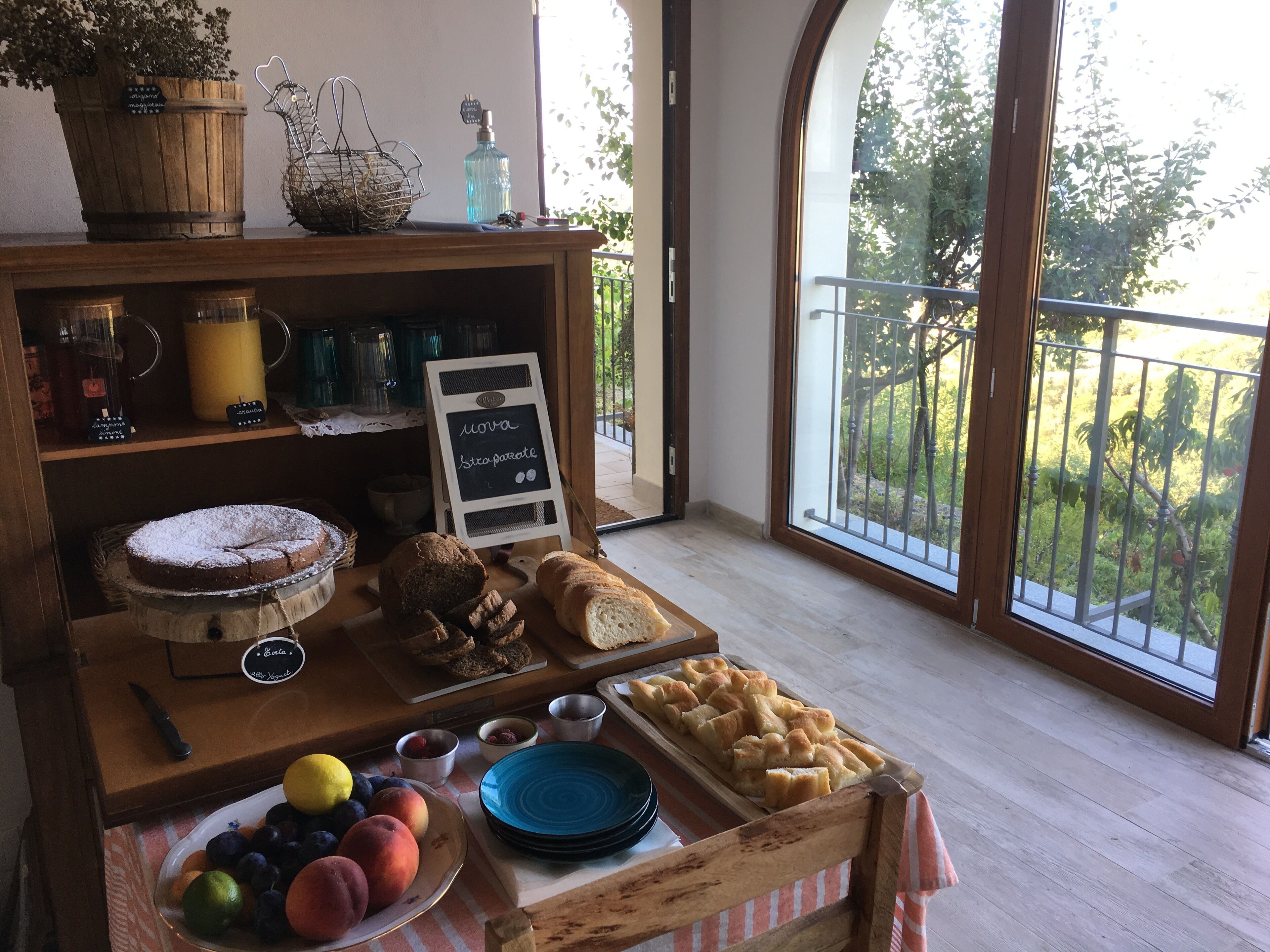 A breakfast buffet with bread, pastries, and fruit on a wooden table, with a sign reading "Moka" and a view of greenery outside.