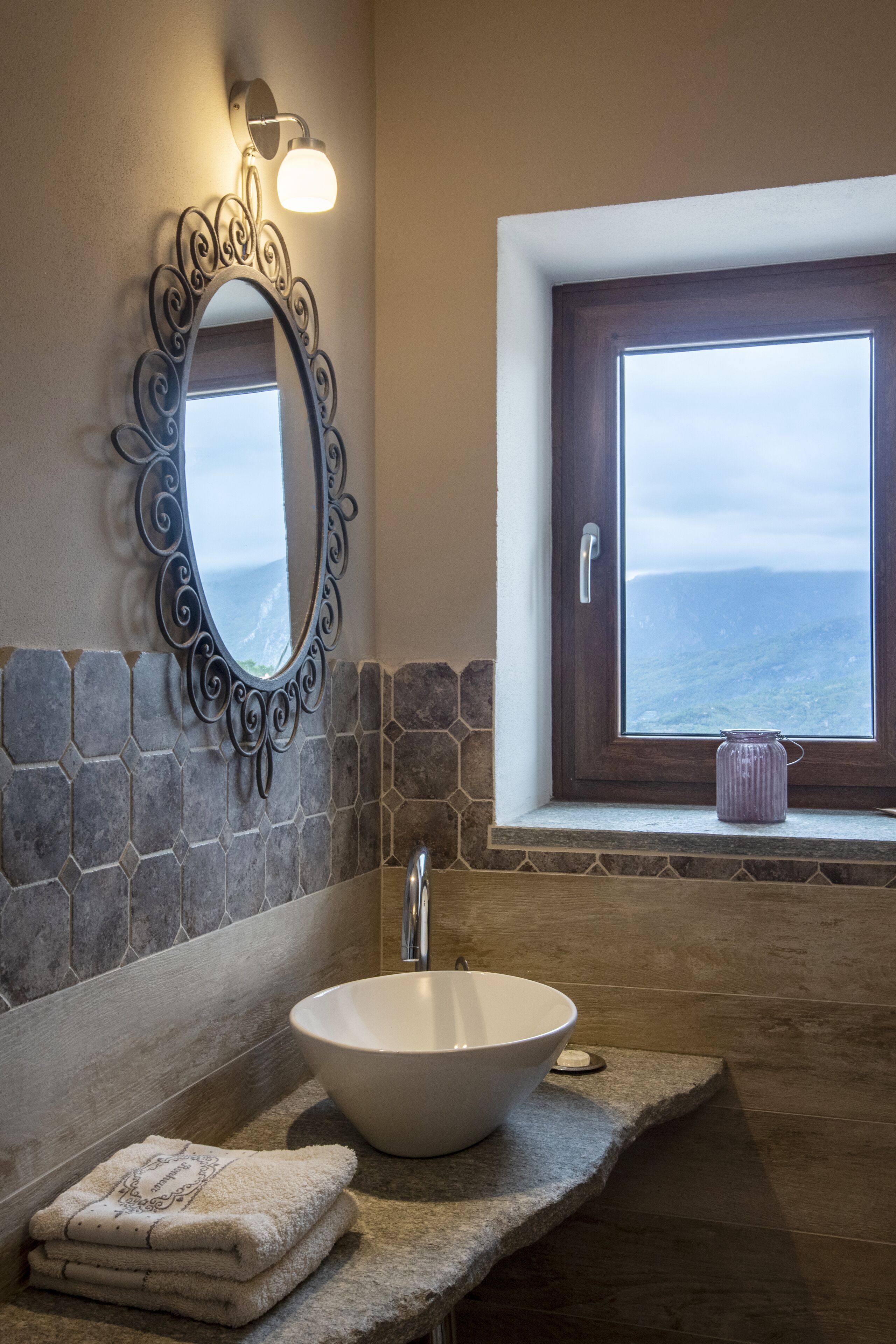 A bathroom with a stone countertop, a white sink, a decorative mirror, and a window with a view of the sea.