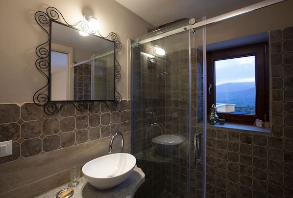 Un baño con pared de piedra, ducha envuelta en vidrio, lavamanos blanco y ventana con vistas.