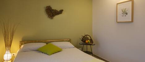 A neatly made bed with a green pillow, a wall-mounted heart decoration, and a framed botanical illustration.