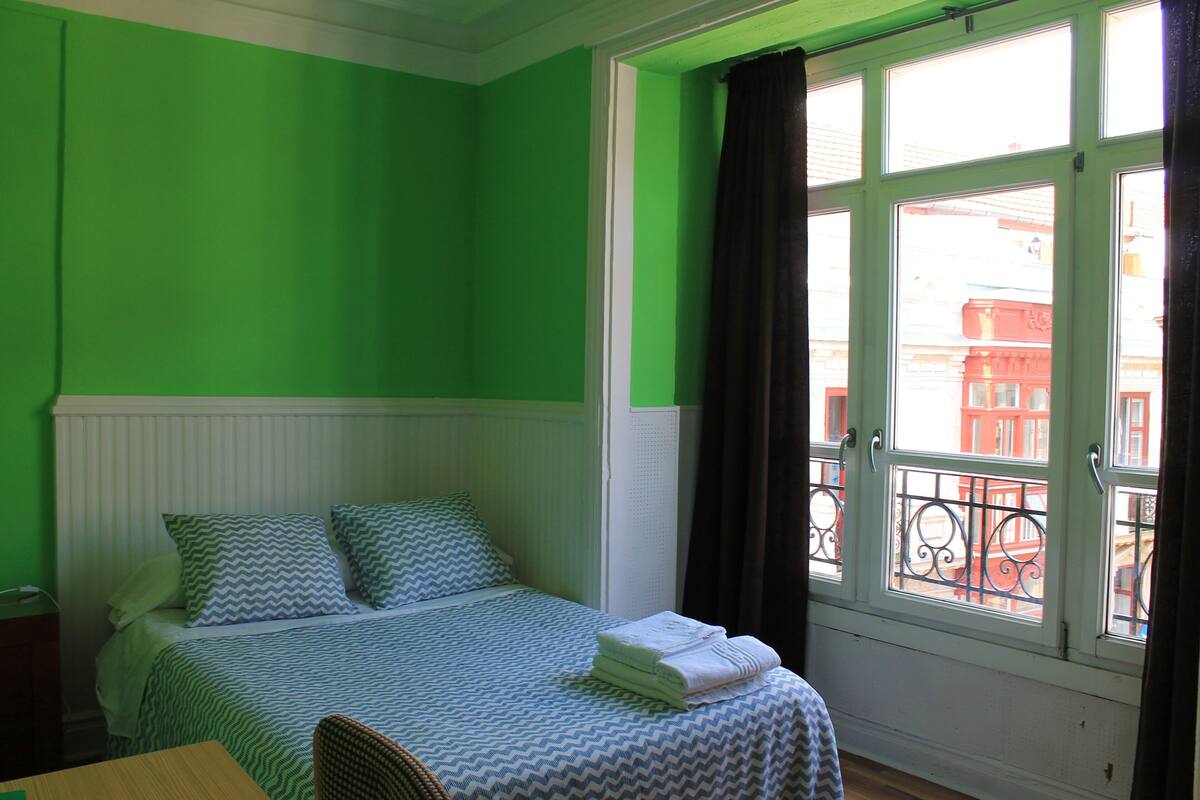 A room with a bed, a window with a view of buildings, and a chair.