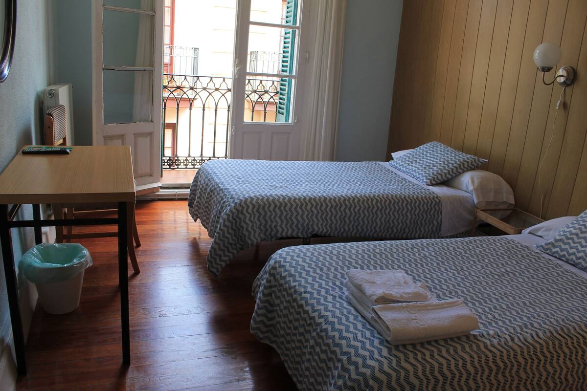 A room with two beds, a wooden table, a trash bin, and a window with a view of the outside.