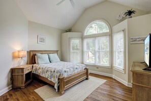 Une chambre à coucher avec un lit en bois, une table de chevet, une lampe, une grande fenêtre avec des stores et un ventilateur de plafond.
