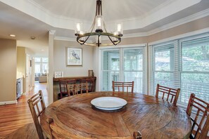 Une salle à manger avec une table ronde en bois, six chaises en bois, un lustre et de grandes fenêtres équipées de stores.