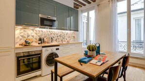 A modern kitchen with a built-in oven, microwave, and washing machine. There is a dining table with chairs and a window with a balcony view. - Sweett | Clery - Two Bedroom Apartment, Sleeps 4 (Paris)