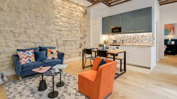 A modern living room with a stone wall, a blue sofa, an orange armchair, and a dining area with a kitchen in the background. - Sweett | Clery - Two Bedroom Apartment, Sleeps 4 (Paris)