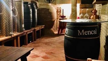 Ein rustikaler Keller mit Steinwänden, Holzbänken, einem großen Fass mit der Aufschrift „Mencal“ und einer rustikalen Kronleuchter.