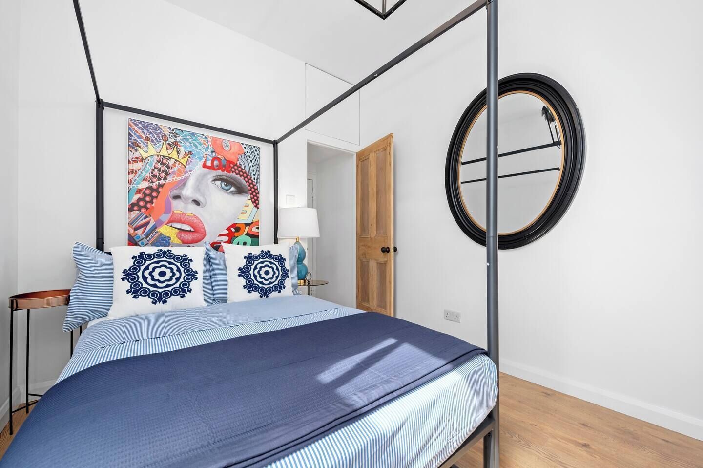 A modern bedroom with a bed, a round mirror, a colorful artwork, and a wooden door.
