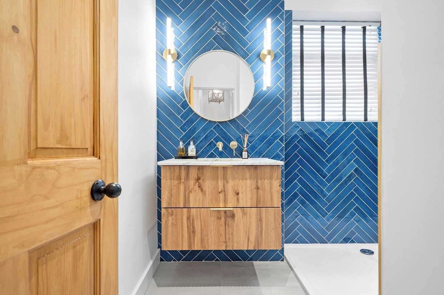 Une salle de bain avec un meuble-lavabo en bois, un miroir rond et des carreaux bleus disposés en motif chevrons.