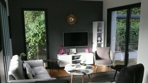 Een moderne woonkamer met een bank, fauteuil en salontafel. Een televisie is aan de muur bevestigd en er is een balkon met uitzicht op groen.