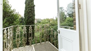 Een balkon met sierlijk metalen hekwerk, uitkijkend op een tuin met een hoge boom en een gebouw op de achtergrond.