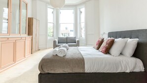 Een slaapkamer met een groot bed, een grijs gestoffeerde fauteuil, een houten garderobe en een groot raam met natuurlijk licht.