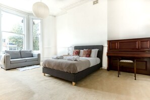Um quarto com uma cama, um sofá cinzento, uma escrivaninha de madeira e uma janela grande.