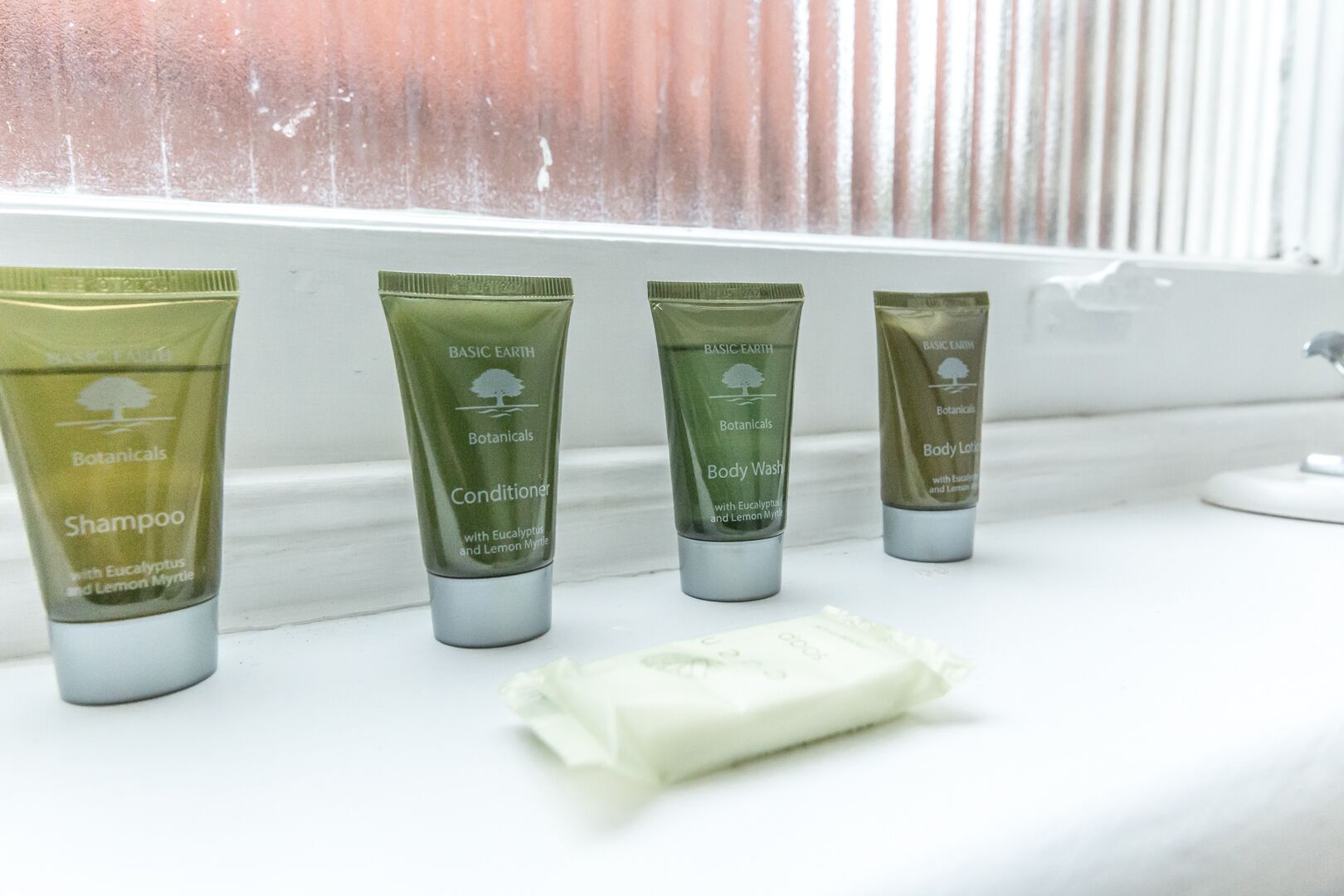 A row of four Basic Earth Botanicals products on a white surface near a window with blinds.