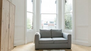 Een kamer met een grijze bank, een zachte witte vloerkleden en grote ramen met uitzicht op gebouwen.