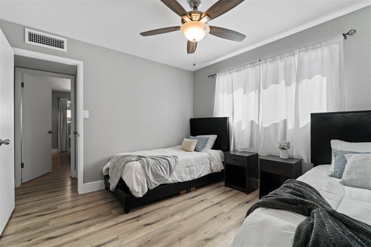 A bedroom with two beds, a ceiling fan, a nightstand, and a window with sheer curtains.