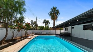 A rectangular swimming pool with clear blue water, surrounded by a white fence and palm trees.