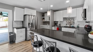 A modern kitchen with white cabinetry, stainless steel appliances, and a black countertop. There are two pendant lights, a built-in oven, and a coffee machine.