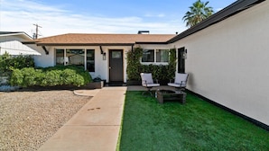 A modern single-story house with a front porch, a small lawn, and two white chairs.
