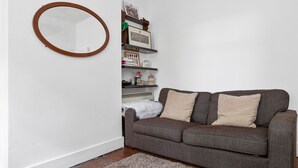 A living room with a grey sofa, two beige cushions, a white rug, a wooden-framed mirror, and a bookshelf with various items. - Cosy 1BR Central Apartment In Bermondsey (London)