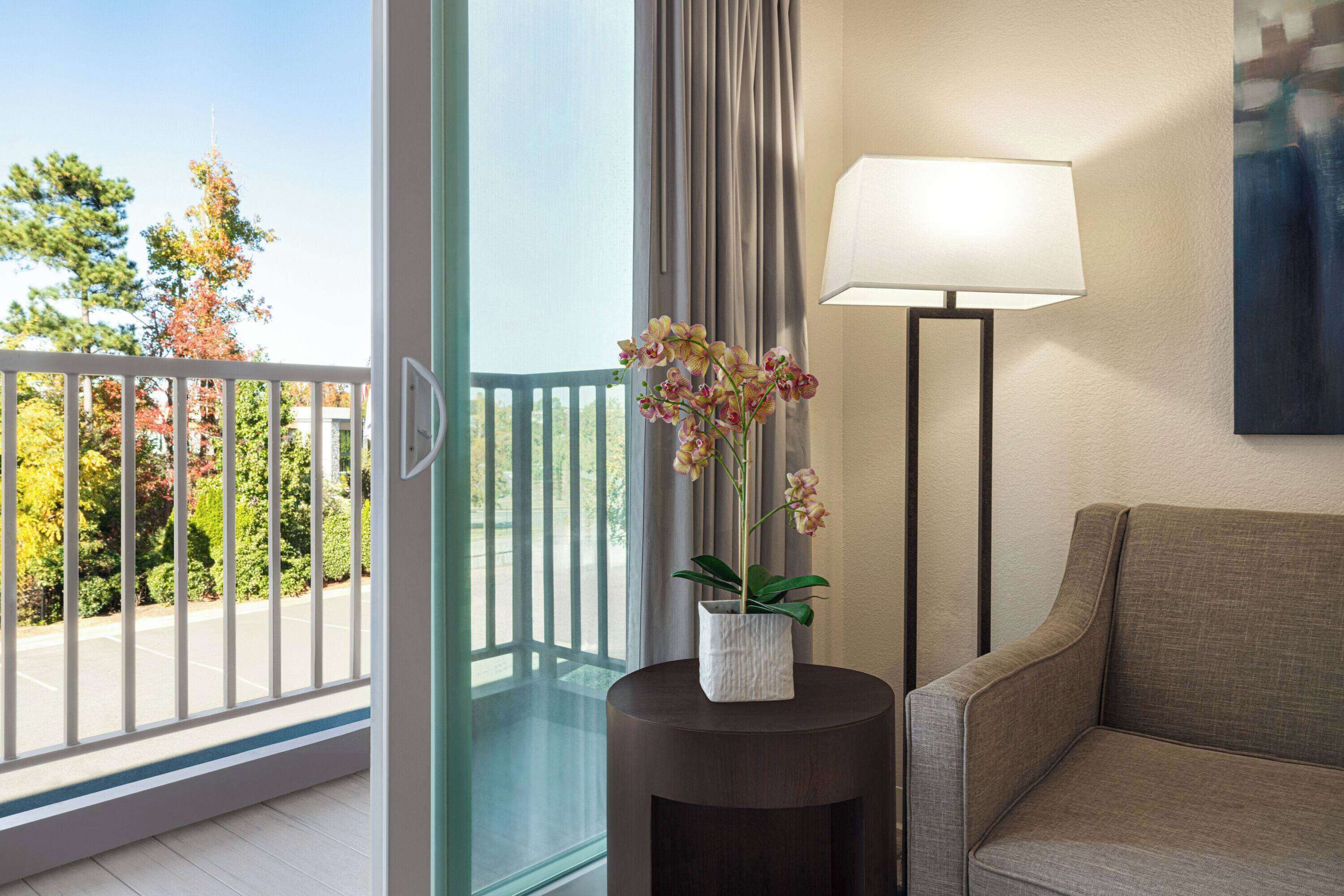 Un balcon donnant sur des arbres et un parking, une table ronde avec une lampe et une orchidée, et un canapé.