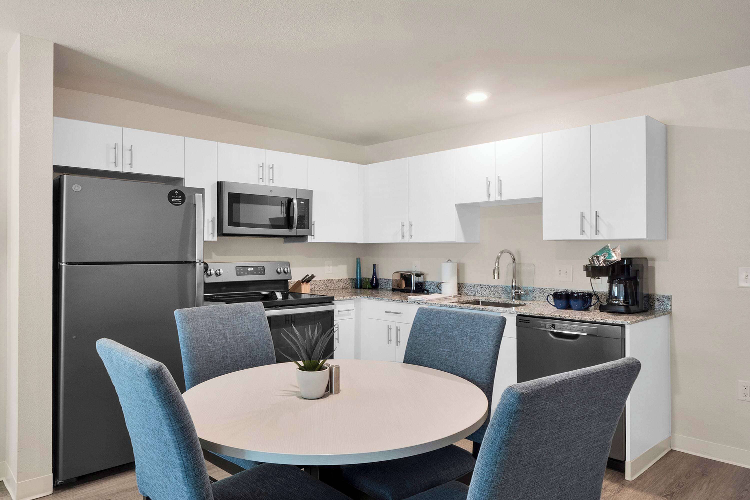 A modern kitchen with white cabinets, a black refrigerator, a microwave, a stove, and a dishwasher. There is a round table with blue chairs and a small potted plant.