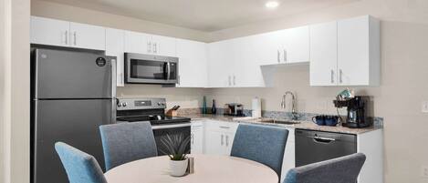 A modern kitchen with white cabinets, a black refrigerator, a microwave, a stove, and a dishwasher. There is a round table with blue chairs and a small potted plant.