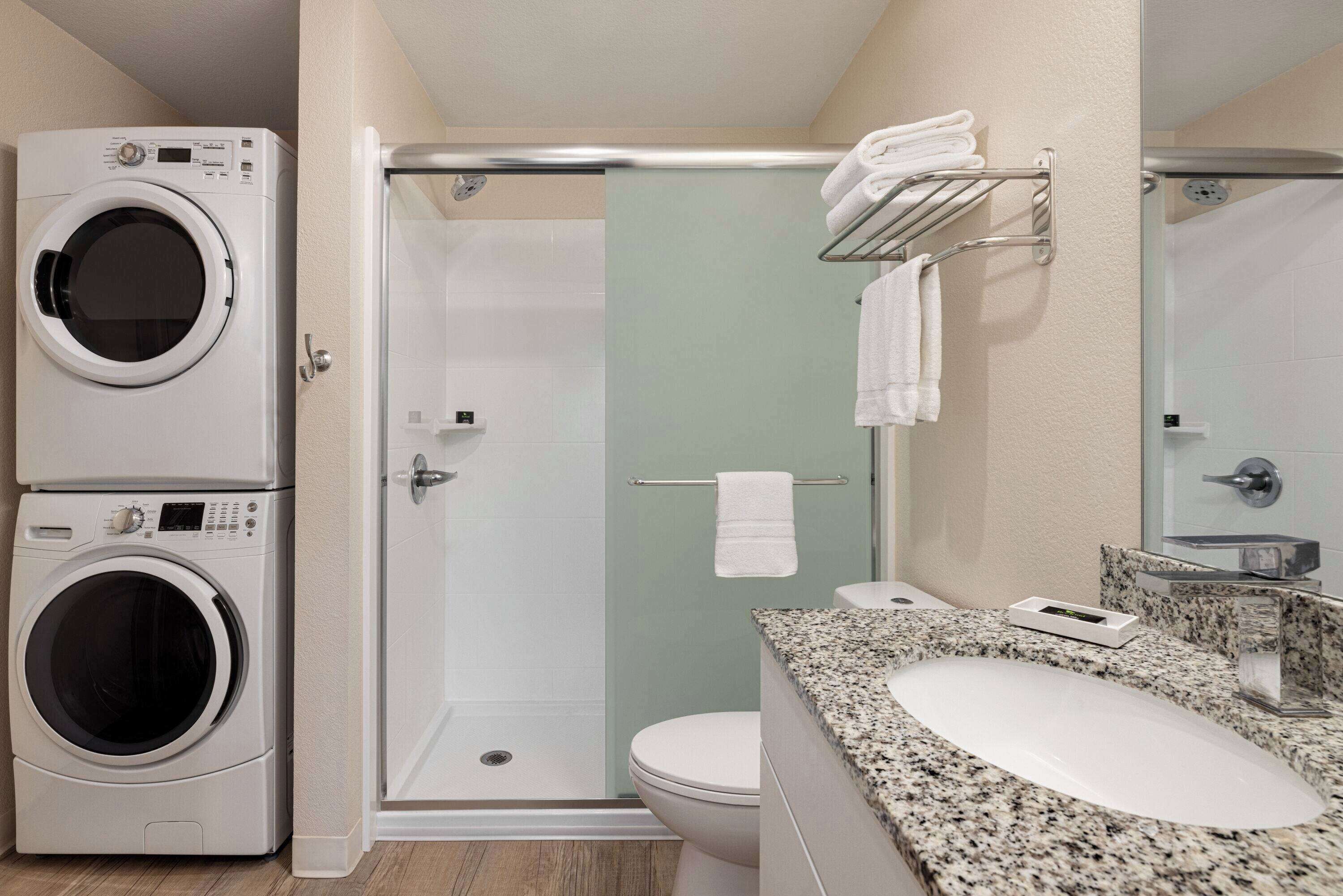 Un baño moderno con lavadora y secadora integradas, cabina de ducha de vidrio y encimera de granito con lavabo.