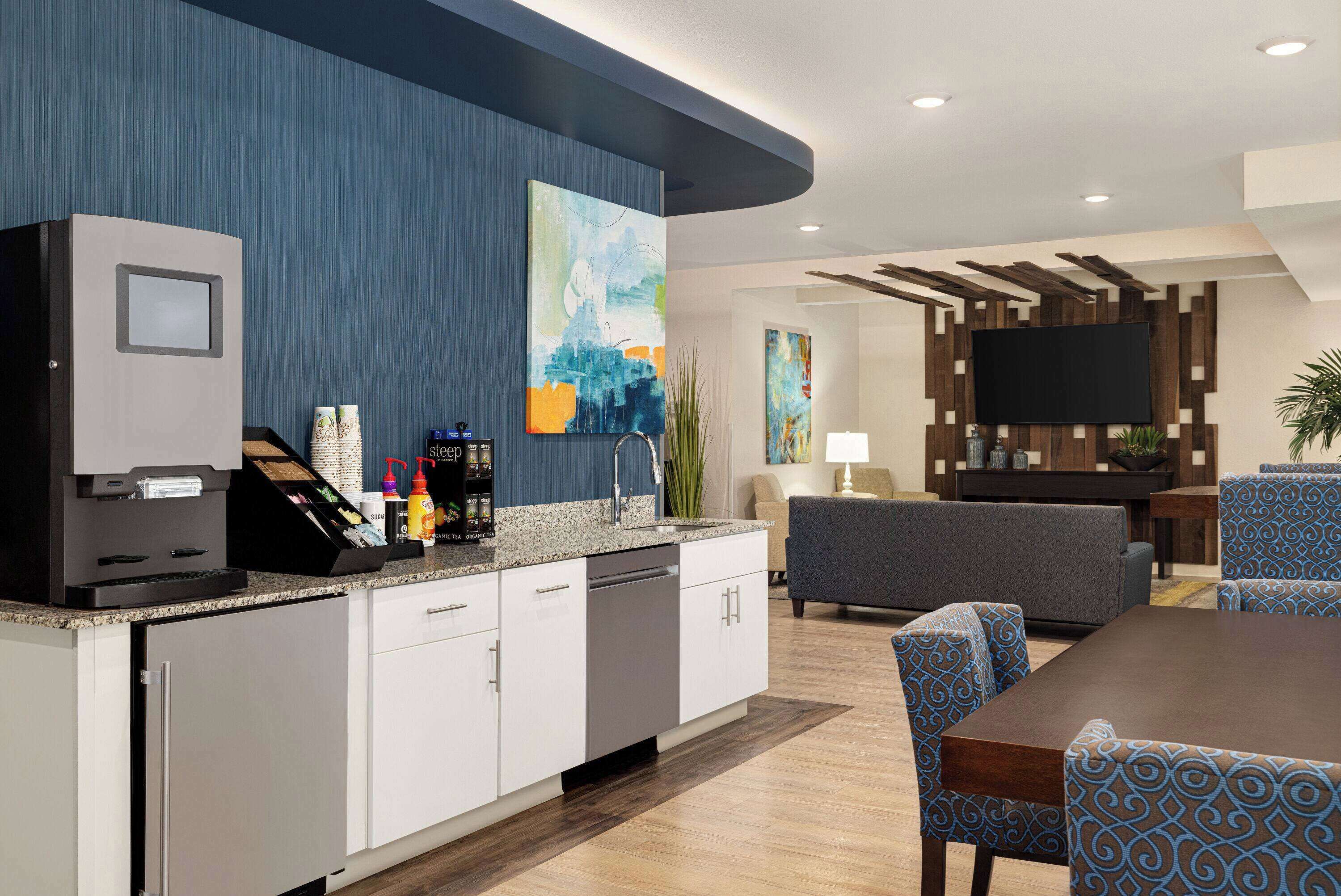 Een moderne keuken met een ingebouwde koffieapparaat, een eethoek met patroonstoelen, een flatscreen-tv bevestigd aan een houten muur, en een bank in de woonkamer.
