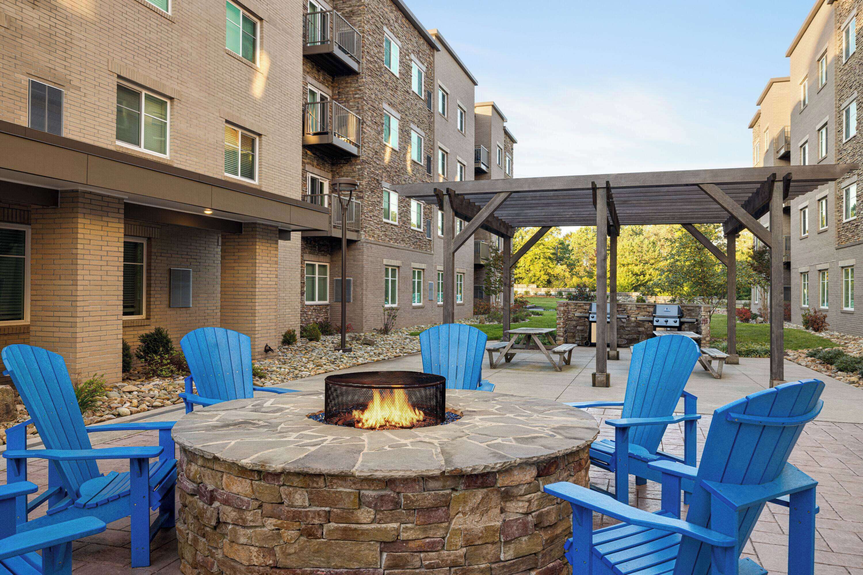 A stone fire pit surrounded by blue Adirondack chairs in an outdoor patio area of a multi-story building with brick walls and balconies.