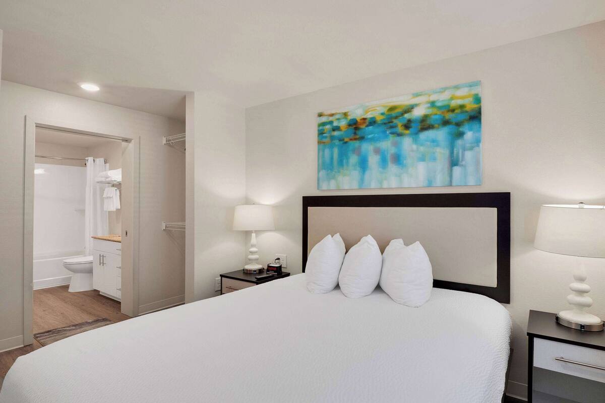 Ein Hotelzimmer mit einem großen Bett, einem Nachttisch mit Lampe, einem Bild an der Wand und einem durch eine offene Tür einsehbarer Badezimmer.