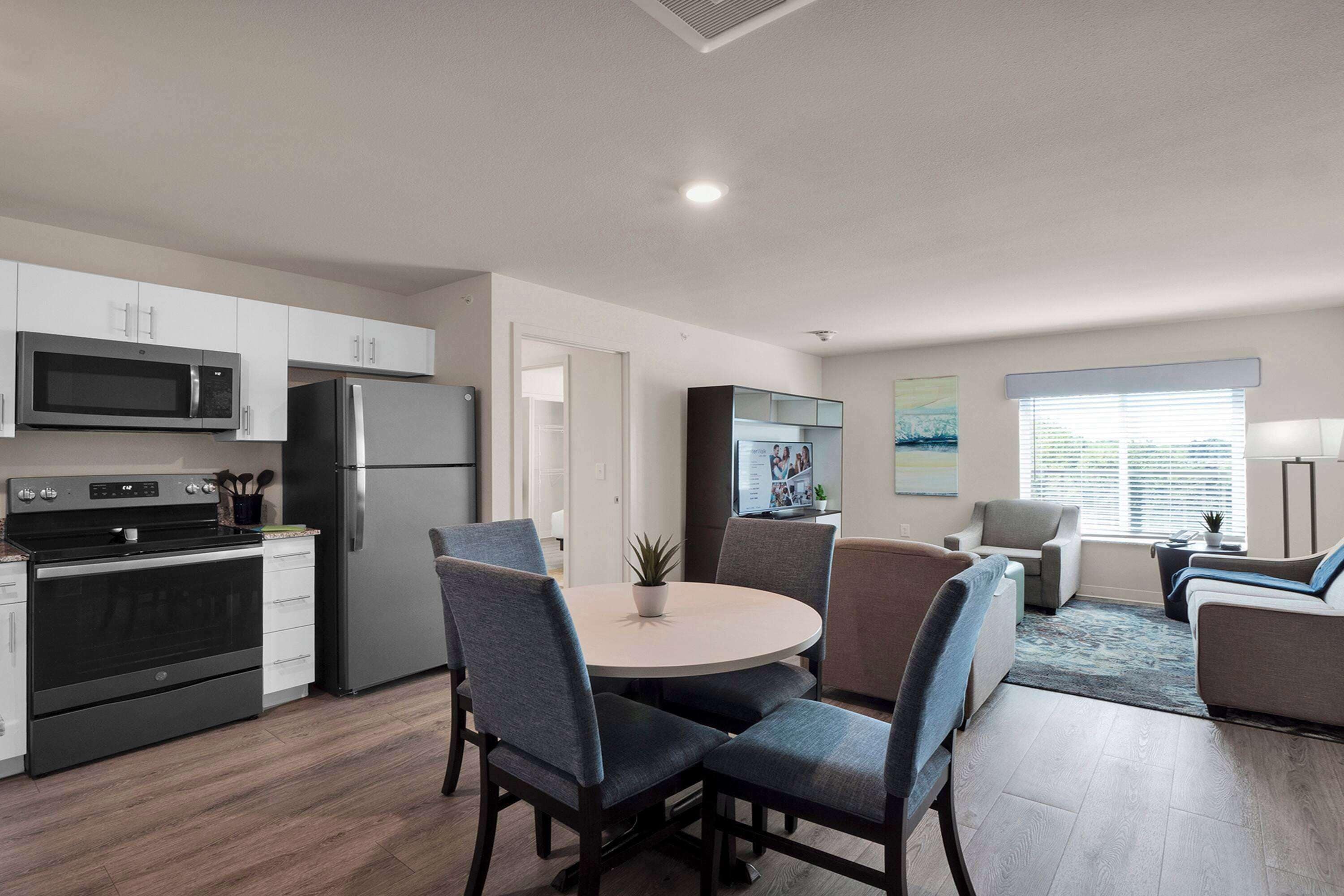 A modern kitchen with stainless steel appliances, a dining area with a round table and chairs, and a living area with a sofa and a TV.