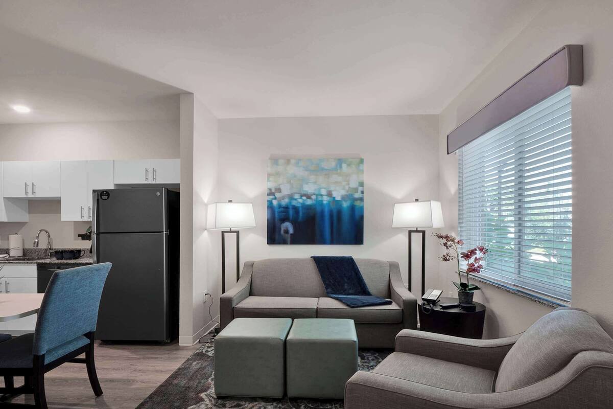 Ein modernes Wohnzimmer mit Sofa, Hocker und Essbereich. Im Hintergrund sind ein Kühlschrank und Küchenschränke zu sehen.
