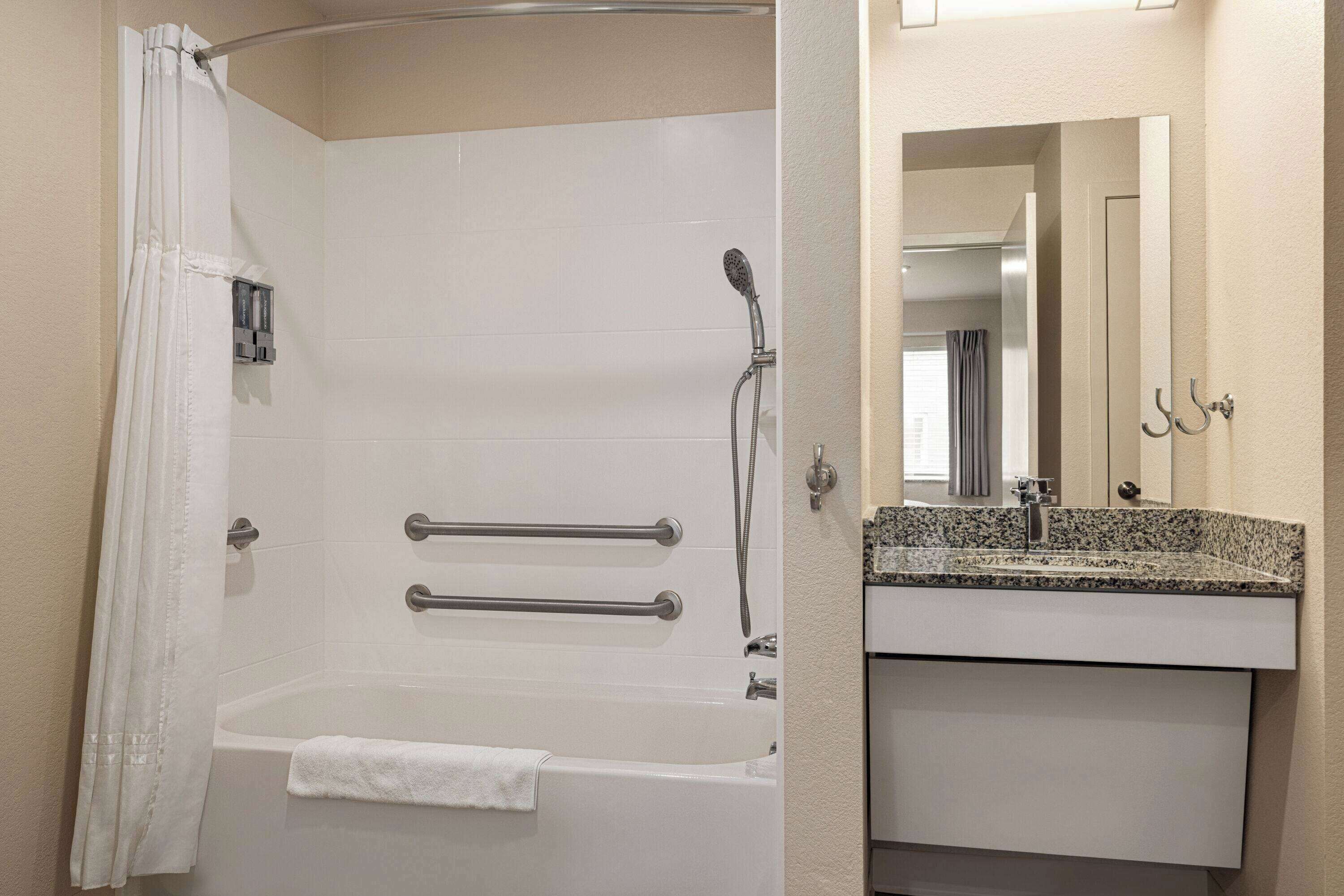 A bathroom with a bathtub, grab bars, a showerhead, a towel, and a sink with a mirror.