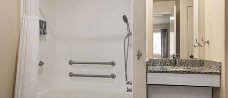 A bathroom with a bathtub, grab bars, a showerhead, a towel, and a sink with a mirror.