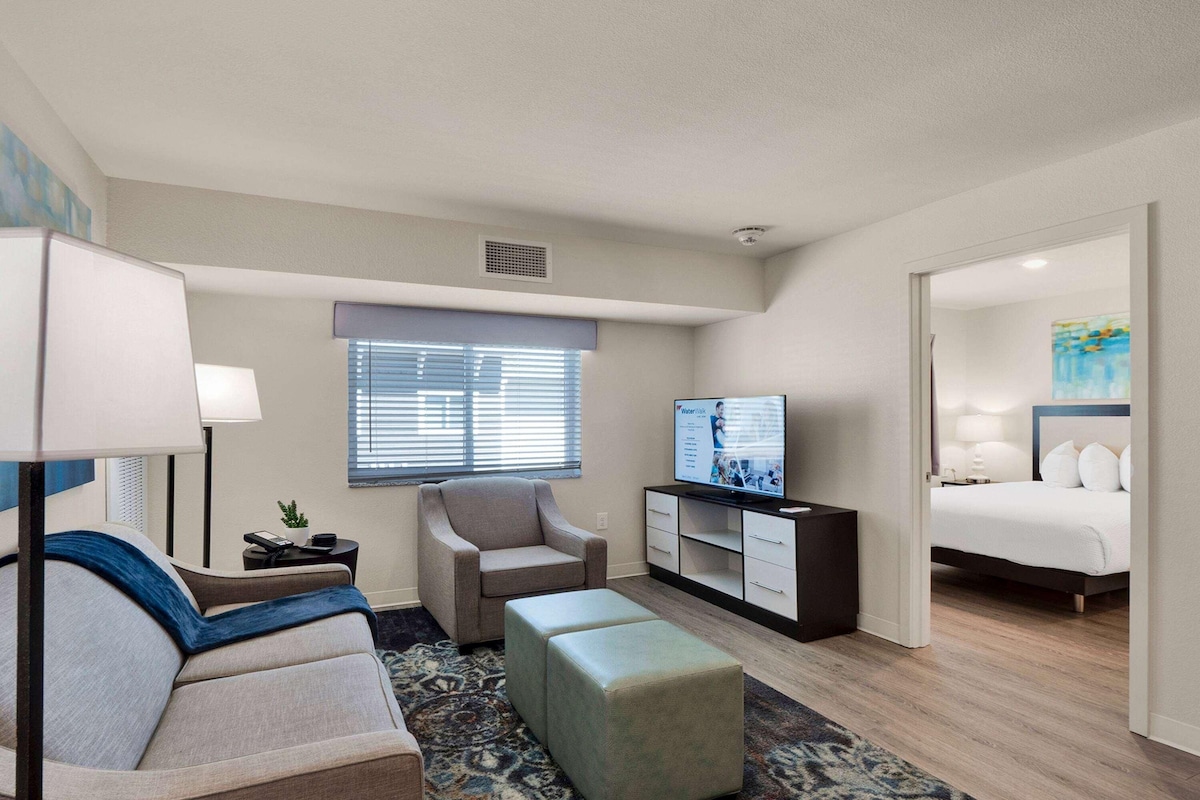 A hotel room with a sofa, armchair, ottoman, TV, and a bed.