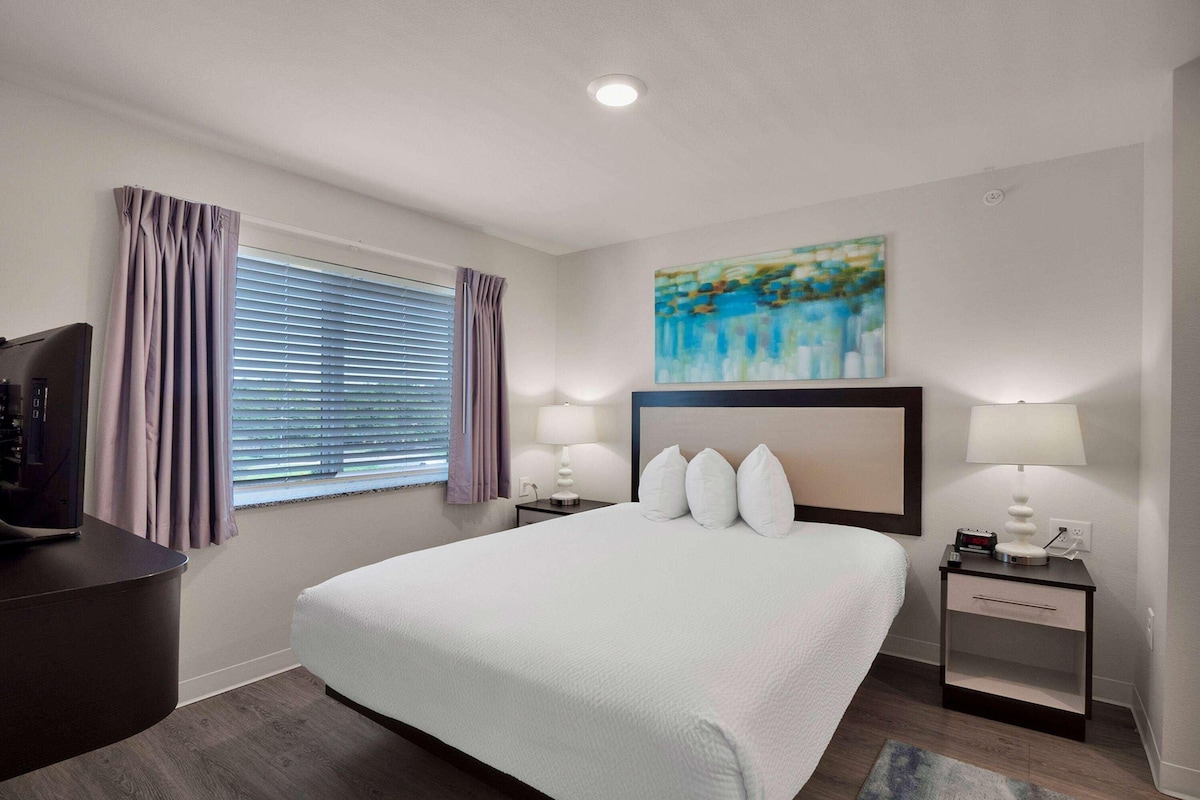 A hotel room with a large bed, two bedside tables with lamps, a painting on the wall, and a window with curtains.