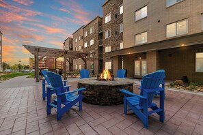 A courtyard with a stone fire pit surrounded by blue Adirondack chairs, a covered seating area, and a brick building with multiple windows. - WaterWalk Extended Stay Raleigh Durham Research Triangle (Morrisville)